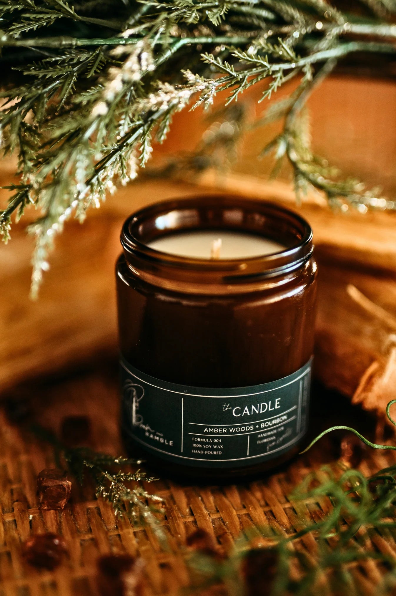 Candle in a brown jar with a green label on a wooden surface with greenery.