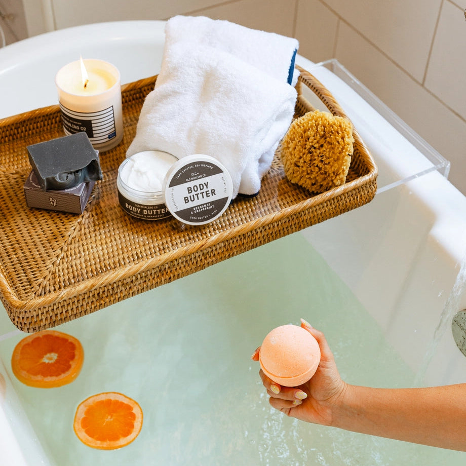 Person holding a pink bath bomb over a bathtub with a tray of bath items including a candle, soap, and loofah.