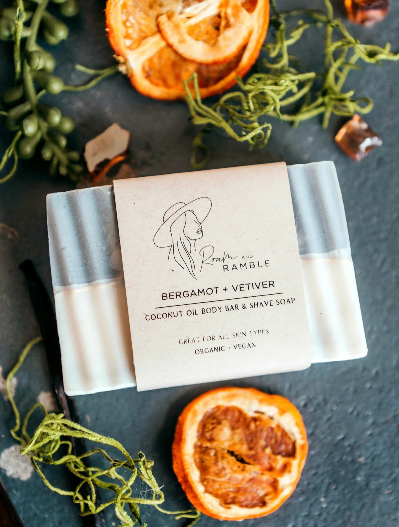 Coconut oil body bar & shave soap with Bergamot + Vetiver scent on a dark surface with dried oranges and greenery.