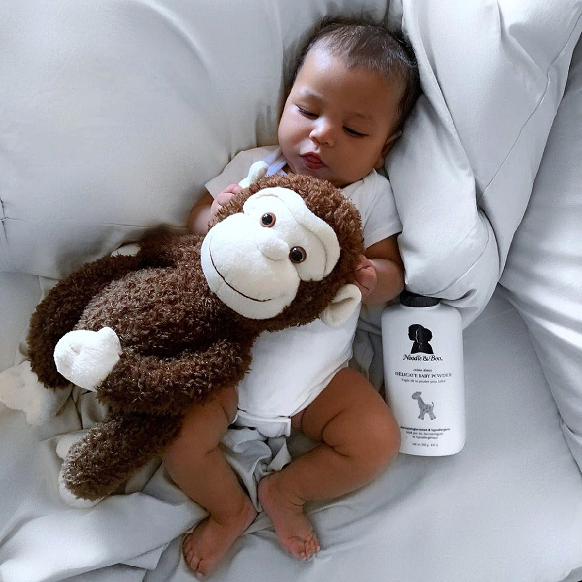 Baby holding a monkey plush toy on a bed with a bottle of Noodle & Boo product next to them.