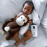 Baby holding a monkey plush toy on a bed with a bottle of Noodle & Boo product next to them.