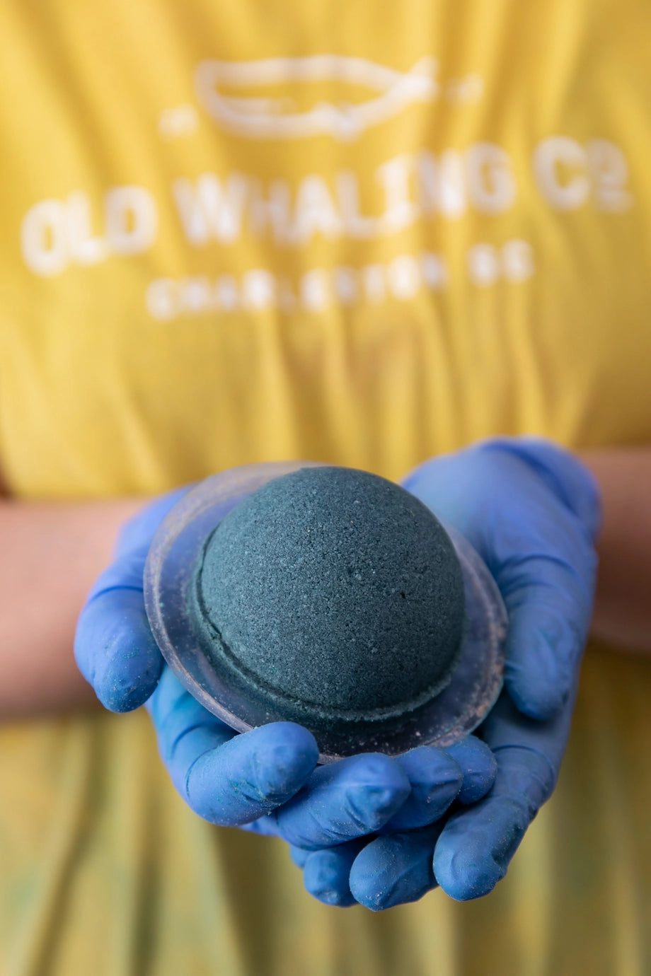 Person wearing a yellow shirt with 'Old Whaling Co' text, holding a blue bath bomb in clear plastic wrap.