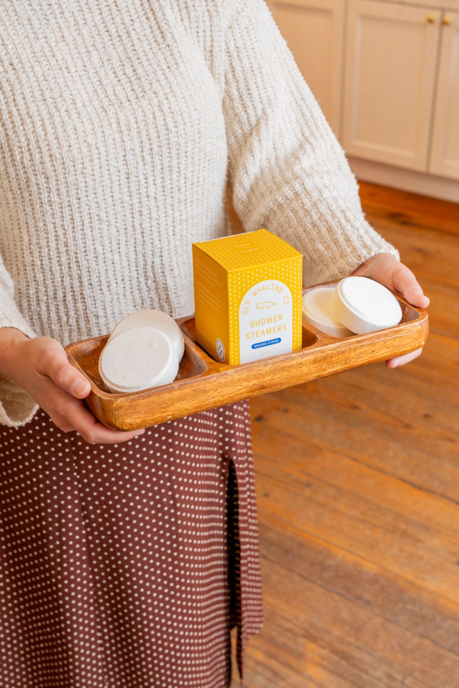 Person holding a wooden tray with skincare products in a kitchen setting